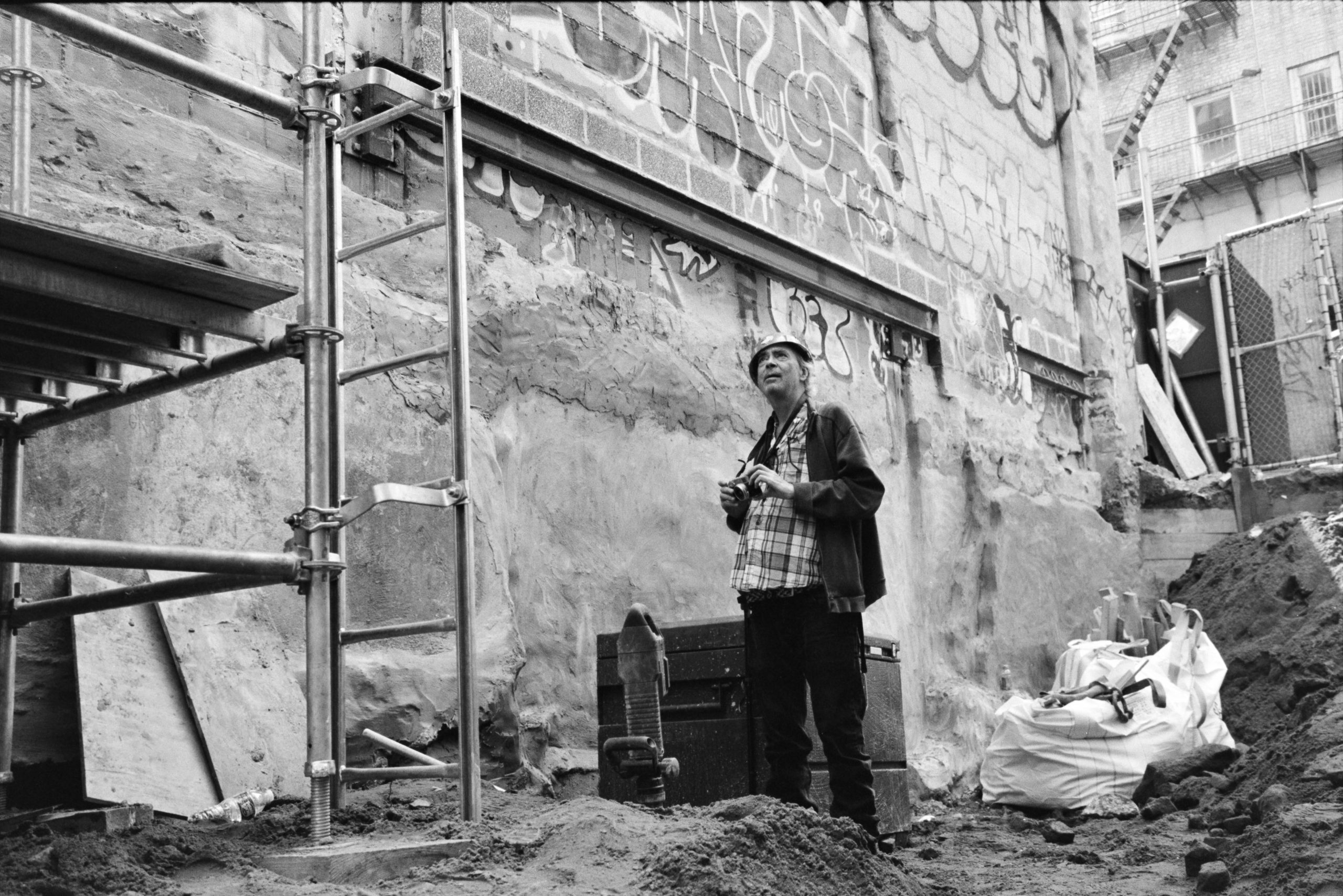 A black and white photograph of man standing in a construction site with a hard hat, clutching a camera at chest height.